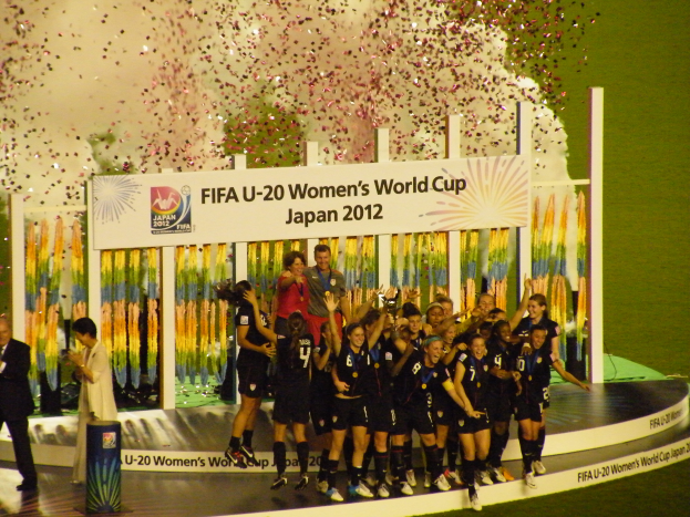 Eine Gruppe von Fussballspielerinnen in schwarzen Uniformen steht auf einer Bühne und feiert ihren Sieg, umgeben von einer Menge mit Kameras, mit einem dekorierten Hintergrund und grasbewachsenem Boden.