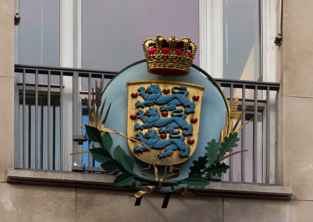 Das Wappen der britischen Königsfamilie, das auf der Außenseite eines Gebäudes dargestellt ist, begleitet von einem Logo mit Krone und Blättern, mit Fenstern und Gittern im Hintergrund.