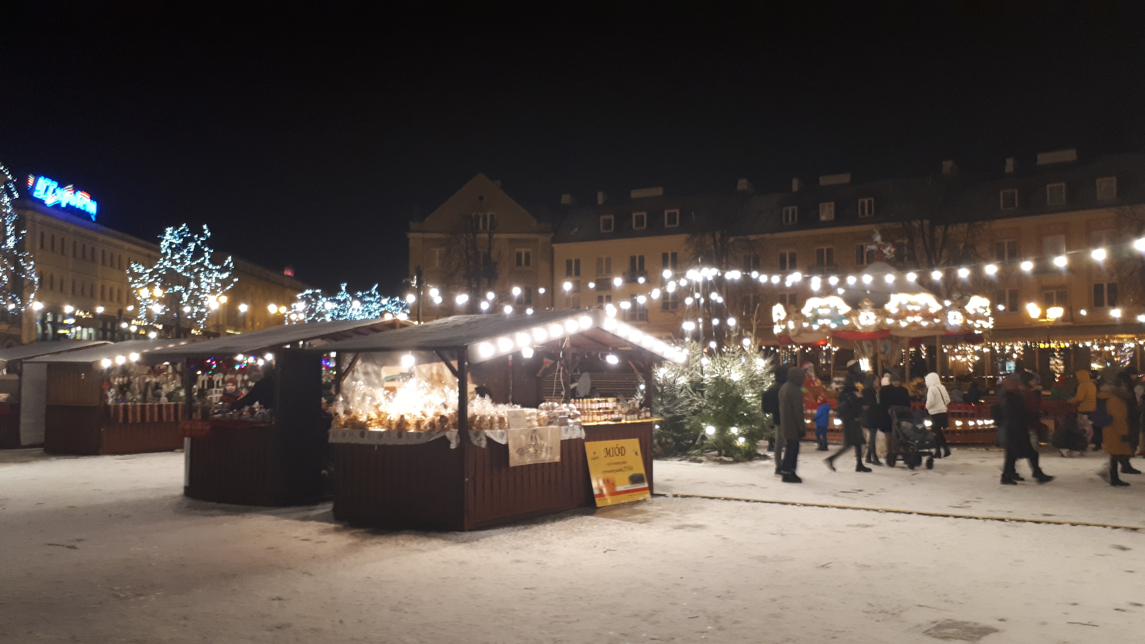 Ein geschäftiges Weihnachtsmarkt auf einer verschneiten Nacht mit Menschen, Ständen mit Lichtern, Pflanzen, Bäumen, Gebäuden, Schildern und einem bewölkten Himmel.