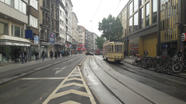Eine Stadtstraße mit einer gelben Tram, Gebäuden mit Fenstern, Schildern, Menschen auf dem Gehweg, parkenden Fahrrädern, einem Straßenschild, Drähten, einem Baum und einem bewölkten Himmel.