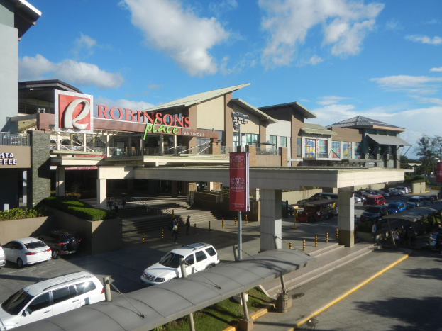 Ein belebtes Einkaufszentrum mit Gebäuden, Geschäften, Fahrzeugen, Fußgängern, Straßenmöbeln, Grünflächen und einer bewölkten Himmel.