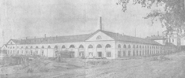 Schwarzes und weißes Foto eines großen, mehrfenstrigen Gebäudes, das von Grün umgeben ist und unter einem klaren Himmel steht, mit dem Text "1892 – das erste Fabrikgebäude der Stadt" unten.