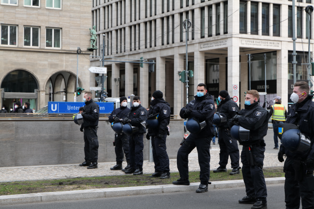 Eine Gruppe von Polizisten in schwarzen Uniformen und Masken steht vor einem Gebäude mit Glasfenstern und Säulen in Berlin, Deutschland, einige halten Helme, mit Laternenmasten, Verkehrszeichen, Schildern mit Text, einer Statue, anderen Menschen und einer grasbewachsenen Fläche im Hintergrund.