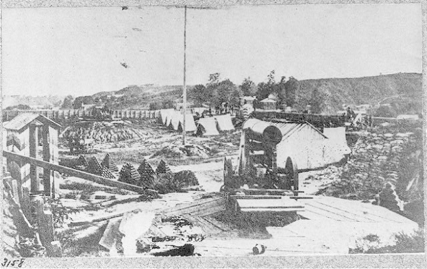 Schwarz-weiß-Foto einer Baustelle mit einer Maschine im Vordergrund, Schuppen, Bäumen, Hügeln und Himmel im Hintergrund, mit Text unten: "Die Bauarbeiten an einer neuen Brücke in der Stadt San Francisco, Kalifornien."