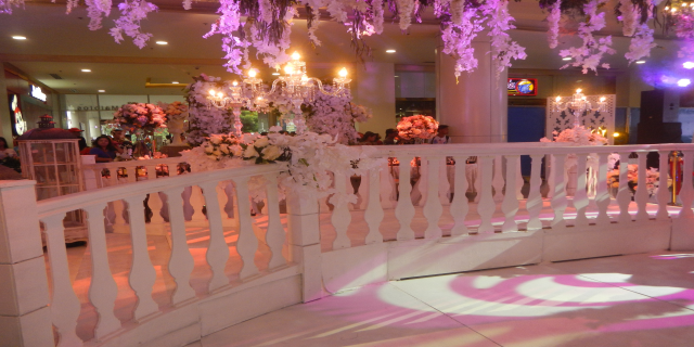 Weißer Handlauf mit Blumen und Kronleuchtern geschmückt, umgeben von Menschen, Säulen und Lichtern im Hintergrund und ein Bildschirm auf der rechten Seite.