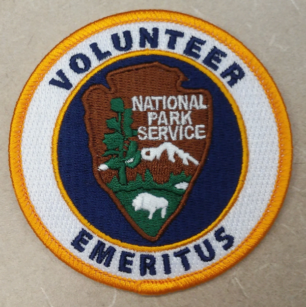 Runder Aufnäher mit blauem Rand und weißem Hintergrund, der den schwarzen Text "Volunteer National Park Service Emeritus" in einem gelben Kreis zeigt.