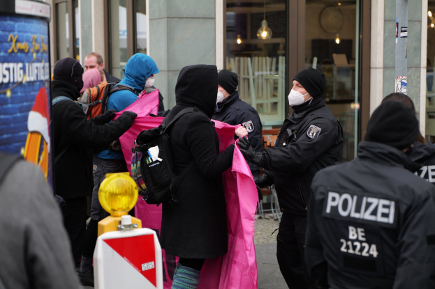 Eine Gruppe von Menschen steht vor einem Glasgebäude in Berlin, wo ein Polizeibeamter eine Frau festnimmt; einige tragen Mützen und tragen Taschen oder Kleidung, und ein Straßenschild ist links zu sehen.