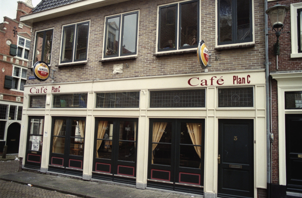 Ein Gebäude mit Fenstern und Türen, ein Schild mit der Aufschrift "Cafe Plan C in Amsterdam, Niederlande", ein Straßenpfahl, eine Baumgruppe und ein bewölkter Himmel.