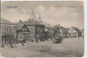 Ein altes Schwarz-Weiß-Foto einer belebten Stadtstraße in Nowogrodeek, Deutschland, mit Gebäuden, Menschen, Karren, Pfählen, Bäumen und Text oben und unten, der besagt "Marktplatz in Nowogrodeek, Deutschland."