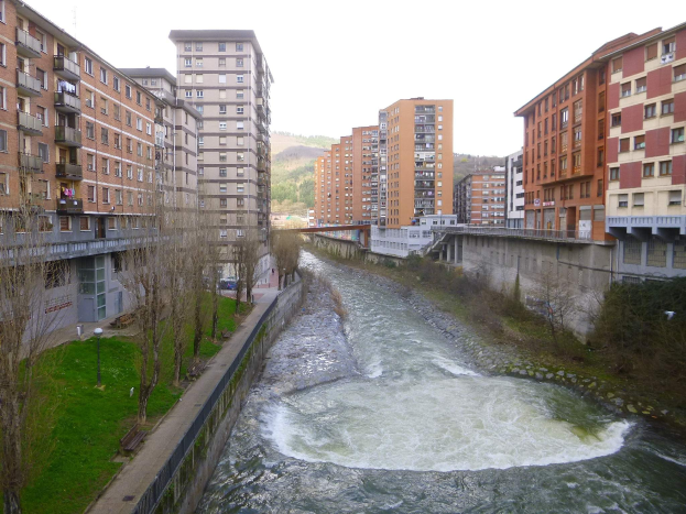 Ein Fluss durchquert eine städtische Gegend mit hohen Gebäuden, gesäumt von grünem Gras, Bäumen und Straßenlaternen, mit einer Brücke, die das Wasser überspannt, und Hügeln in der Ferne unter einem klaren blauen Himmel.