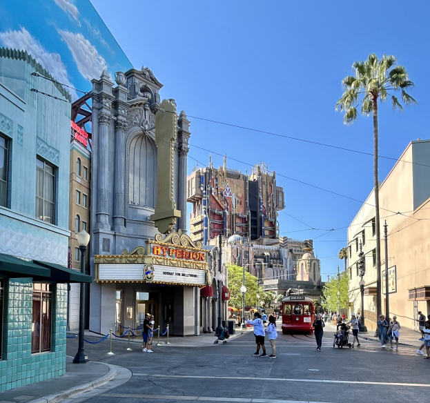 Eine belebte StraßenSzene auf dem Universal Studios Hollywood mit Passanten, einem Fahrzeug, Bäumen, Straßenlaternen, Schildern und einem Gebäude im Hintergrund unter einem sichtbaren Himmel.