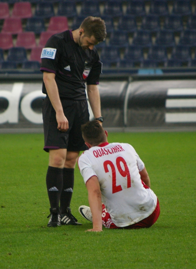 Ein Fussballspieler in Sportkleidung sitzt neben einem Schiedsrichter in einer Stadionumgebung, wobei im Hintergrund Bretter und Stühle zu sehen sind.