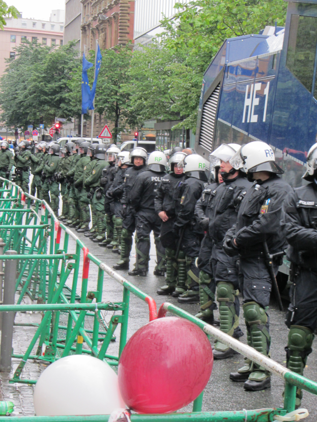 Eine Gruppe von Polizisten in Helmen und mit Waffen vor einer Absperrung auf einer Straße mit Gelöndern und Luftballons, mit Bäumen, Gebäuden, Flaggen und einem klaren blauen Himmel im Hintergrund.