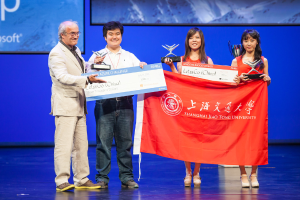 Vier Personen stehen auf einer Bühne, jede hält Auszeichnungen, Schecks und eine Flagge, sie lächeln mit einem digitalen Bildschirm im Hintergrund, der eine Projektion anzeigt.