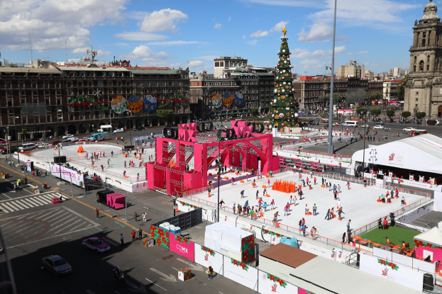 Eine große Gruppe von Menschen auf einer Eisbahn in einer Stadt, umgeben von Gebäuden, Bäumen, Polen, Lichtern und Fahrzeugen auf der Straße, mit einem Weihnachtsbaum in der Mitte und einem bewölkten Himmel.