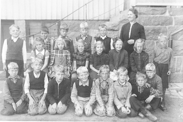 Schwarzes und weißes Foto einer Gruppe von Kindern, die vor einem Gebäude mit Treppe und Geländer posieren, mit der Beschriftung "1930er Schulkinder" unten.
