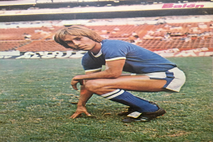 Ein Mann kniet auf einem Fuballfeld umgeben von Menschen, die auf Stühlen und Brettern mit Text sitzen.