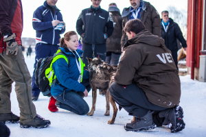 Eine Gruppe von Menschen, einige sitzen und einige stehen, mit einem Hund, alle in einer eisigen Umgebung.