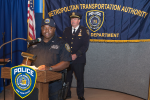 Zwei Polizeibeamte stehen vor einem Rednerpult, einer Tafel mit Symbolen und Text, einer Fahne und einem Tisch, wobei einer der Beamten Papiere hölt und in ein Mikrofon spricht.