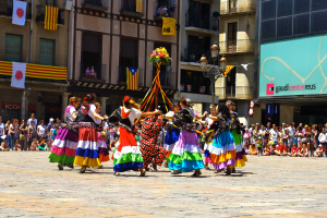 Eine Gruppe von Frauen in bunten Kleidern, die auf der Straße tanzen, umgeben von einer Menschenmenge, mit Gebäuden, Geländern, Balkonen, Fahnen, einem Laternenmast und einer Tafel mit Aufschrift im Hintergrund.