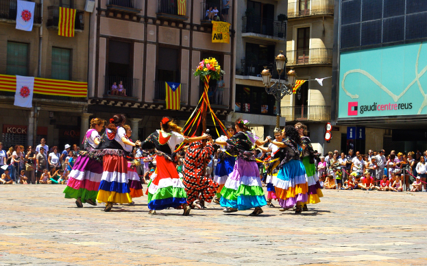 Eine Gruppe von Frauen in bunten Kleidern, die auf der Straße tanzen, umgeben von einer Menschenmenge, mit Gebäuden, Geländern, Balkonen, Fahnen, einem Laternenmast und einer Tafel mit Aufschrift im Hintergrund.