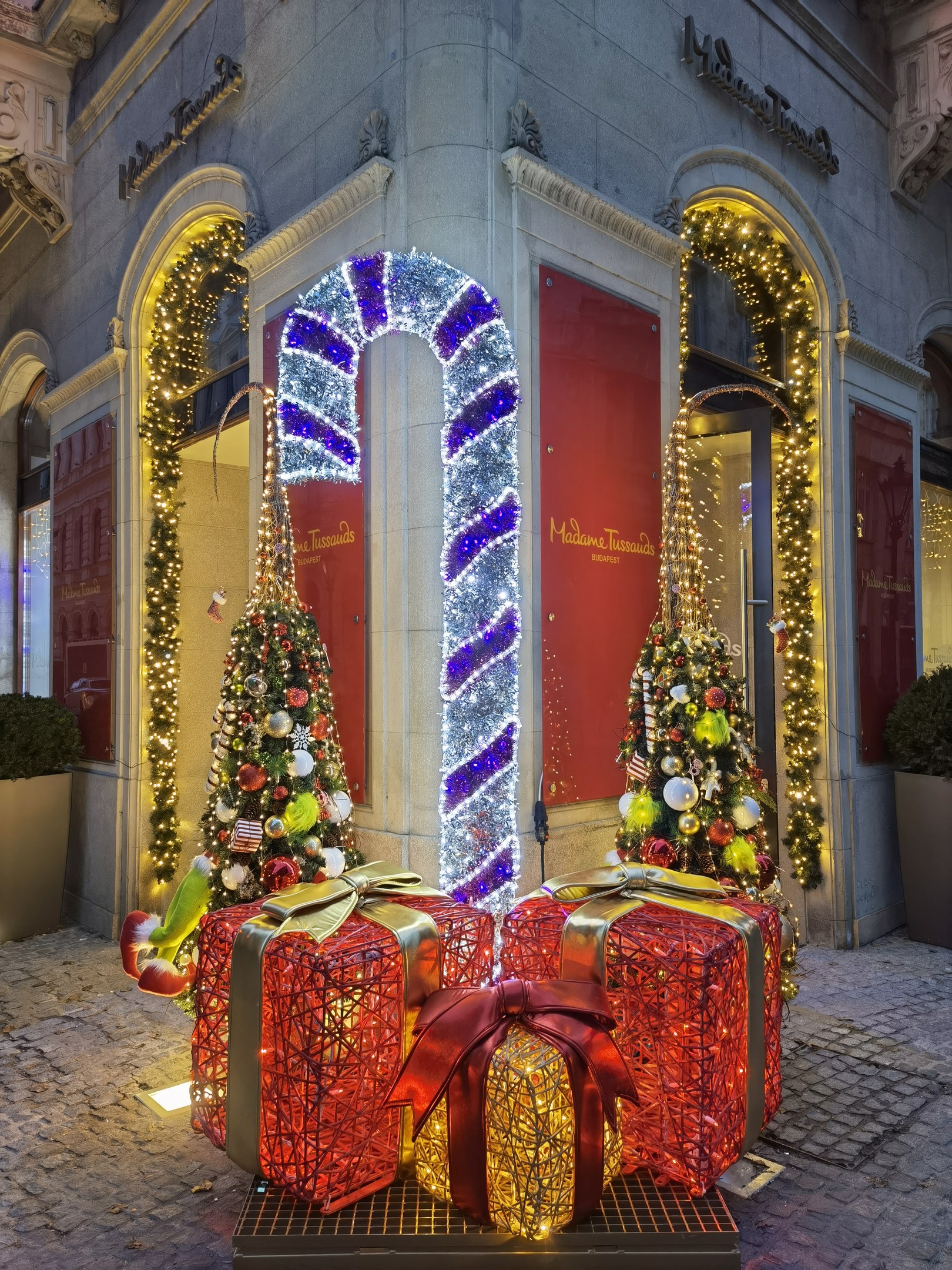 Ein Gebäude mit festlicher Weihnachtsdekoration, das zwei geschmückte Tannenbäume, Geschenkschachteln, Topfpflanzen, eine Tür und Fenster zeigt.