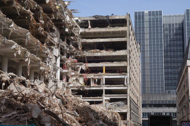 Ein großes Gebäude wird in der Stadtmitte abgerissen, umgeben von intakten Gebäuden, Straßeninfrastruktur, Fahrzeugen, Bäumen und einem klaren Himmel.