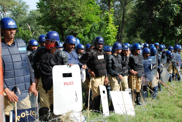Gruppe von Polizisten in Helmen, Schutzwesten und mit Schilden und Schlagstöcken vor einem Stacheldrahtzaun, mit grasbewachsenem Boden, Bäumen und einem klaren blauen Himmel im Hintergrund.