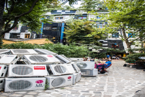 Luftkühler sind links unten auf dem Boden platziert, mit einem Weg nach rechts, Menschen auf Stühlen neben dem Weg und Gebäuden, Bäumen und Pflanzen oben.