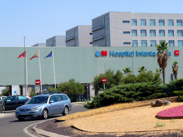 Krankenhaus mit parkenden Autos, umgeben von städtischen Elementen wie Gebäuden, Straßeninfrastruktur, Fahrzeugen, Grünflächen und einem klaren Himmel.