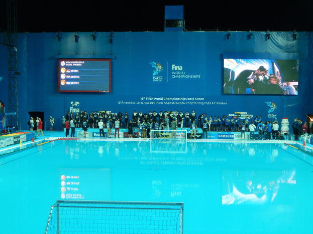 Gruppe von Menschen um ein Hallenbad mit Netz am Boden herumstehend, mit einer Wand im Hintergrund, die "FINA World Championships 2015 Kazan"-Text und Logos anzeigt.