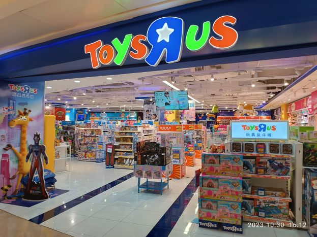 Ein Toys R Us Geschäft in einem Einkaufszentrum, mit Regalen voller Spielzeug und anderer Gegenstände, einem Namensschild mit Text, Lampen an der Decke und einem Wasserzeichen in der rechten unteren Ecke.