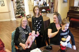 Ein Raum mit einer Wand, die ein Foto Display, einen Weihnachtsbaum, eine Lampe und Blumenpflanzen zeigt; Treppen führen nach oben und einige Menschen sitzen an Tischen mit einem Spielzeug und einer Kamera auf einem davon.