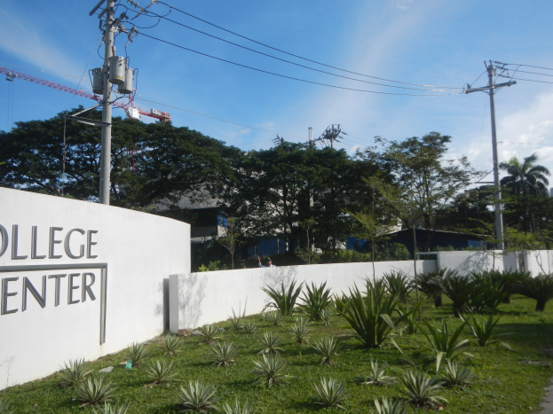 College Center-Eingangsschild an einer Gebäudewand umgeben von Grün, einem Weg, Strommasten, Bäumen, fernen Gebäuden, einem Kran und einem bewölkten Himmel.
