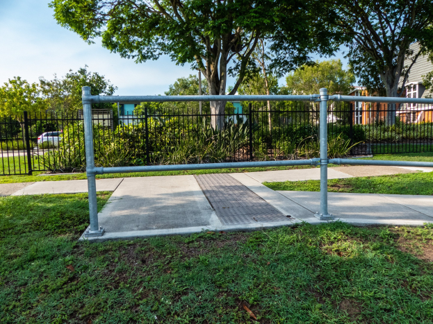 Metallzaun in einer grasigen Gegend mit umliegenden Pflanzen, Bäumen, einem Auto auf der Straße, Gebäuden im Hintergrund und einem klaren blauen Himmel.