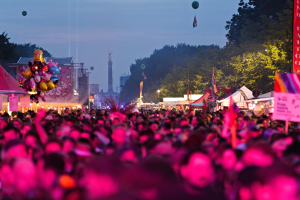 Eine gro%C3%9Fe Menge auf einem nächtlichen Musikfestival mit bunten Lichtern, Ballons, Zelten, Bannern, Fahnen, Bäumen, einem Turm und einem sternenklaren Himmel im Hintergrund.