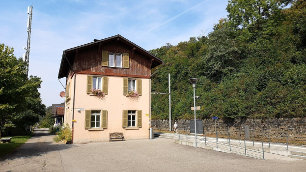 Ferienhaus zum Verkauf in Baden-Württemberg, Deutschland, mit einem mehrfenstrigen Gebäude mit einer vorderen Einfriedung, umgeben von Straßen-Elementen und Grün unter einem bewölkten Himmel.