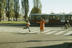 Eine Frau und ein Kind überqueren die Straße vor einem Bus, mit einem Radfahrer auf der rechten Seite der Straße, Bäumen, Fahrzeugen, Gebäuden und einem klaren blauen Himmel im Hintergrund.