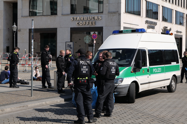 Polizisten vor einem Starbucks-Café mit einem Van auf der rechten Seite und Menschen auf der linken Seite, vor einem Hintergrund aus einem Gebäude, einem Schild, einem Laternenpfahl und einem Zaun.