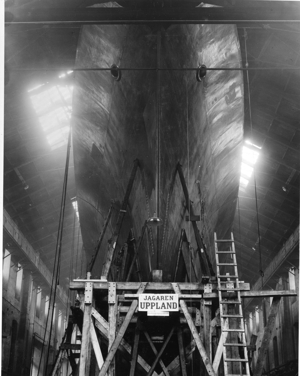 Schwarzes und weißes Foto einer großen Boot im Bau in einer Werft, mit einer Leiter, Holzpfosten, einem Schild, Metallrahmen, Deckenleuchten und Fenstern.