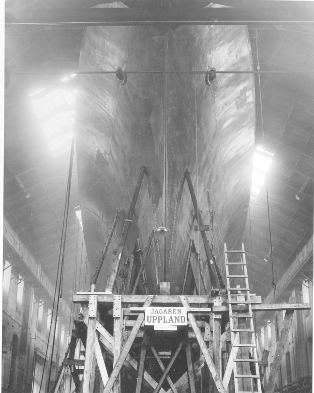 Schwarzes und weißes Foto einer großen Boot im Bau in einer Werft, mit einer Leiter, Holzpfosten, einem Schild, Metallrahmen, Deckenleuchten und Fenstern.