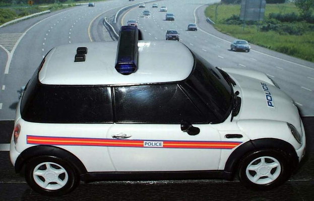 Weißer Polizeiwagen mit "Polizei"-Aufschrift auf einer Landstraße außerhalb der Stadt, umgeben von Verkehr, Gras und Bäumen auf beiden Seiten.