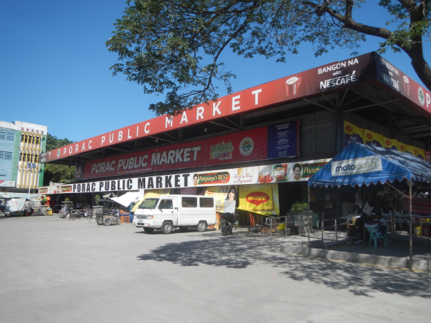 Ein ├Âffentlicher Markt mit einem vor ihm geparkten Van, umgeben von Geb├Ąuden, Gesch├Ąften, Schildern, Kraftfahrzeugen auf der Stra├če, Menschen unter einem Zelt sitzend, Gel├Ąndern, B├Ąumen und einem klaren blauen Himmel.