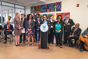 Eine Gruppe von Menschen, die vor einem Podium mit Mikrofon und einer Tafel mit Text darauf stehen und sitzen, mit Flaggen, einer Tafel, einem Feuerlöscher und Deckenleuchten an der Rückwand.