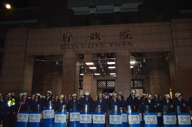 Eine Gruppe von Polizisten in Helmen und Schilden steht vor einem Gebäude mit Text an der Wand und Deckenleuchten, mit anderen Gebäuden im Hintergrund.
