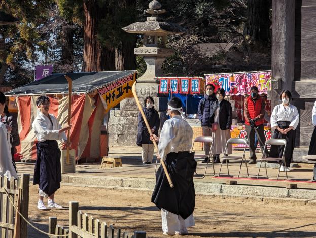 Eine Gruppe von Menschen in traditioneller Kleidung steht im Freien in Kyoto, einige tragen Masken und halten Holzstöcke, mit Stühlen, Bannern und einem Zelt im Hintergrund unter einem klaren blauen Himmel.