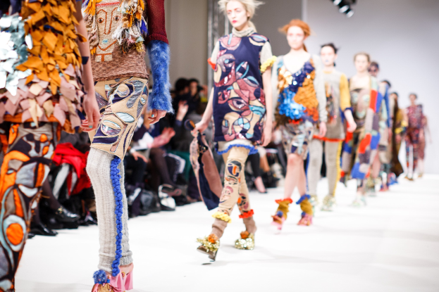 Eine Gruppe Von Frauen, Die Eine Laufsteg In London Fashion Week Heruntergehen, Mit Einer Zuschauerschaft Im Hintergrund Unter Helleren Lichtern.