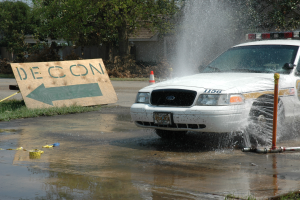 Ein Polizeiwagen fährt durch eine Wasserlache, mit einer Tafel mit Text, einem Verkehrskegel und anderen Gegenständen auf dem Boden, mit Bäumen, Gebäuden und einem klaren blauen Himmel im Hintergrund.