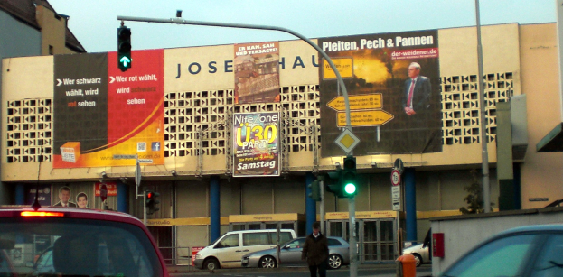 Ein rotes Auto fährt auf einer Straße neben einer Ampel vorbei, mit einer Person, die auf dem Gehweg im Vordergrund läuft; im Hintergrund sind Gebäude, Pfosten, Verkehrszeichen, Werbetafeln und Reklamen sowie ein klarer blauer Himmel zu sehen.
