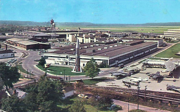 Altes Schwarz-Weiß-Foto eines Flughafens mit parkenden Flugzeugen, Gebäuden, Bäumen, Straßen, Fahrzeugen, Strommasten, Gras, Eisenbahnschienen, Türmen, Hügeln und einem klaren Himmel, mit Text am unteren Rand.